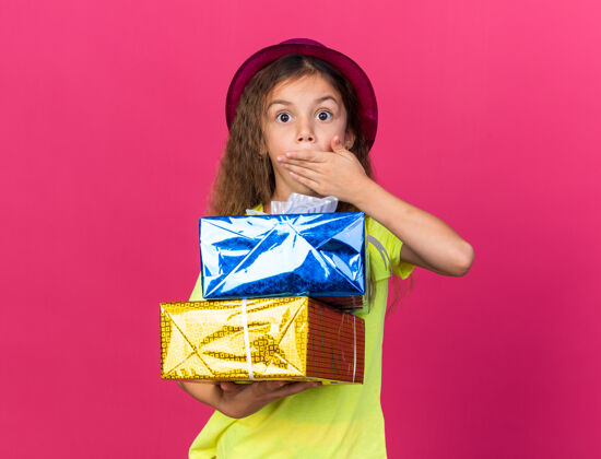 惊喜惊讶的白人小女孩 戴着紫色派对帽 手放在嘴上 手里拿着礼品盒 孤立地放在粉色的墙上 还有复制空间小帽子白种人