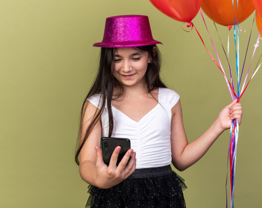 年轻年轻的白人女孩戴着紫色的派对帽看着手机 手里拿着氦气球 孤立地站在橄榄绿的墙上 留着复印空间孤立请橄榄