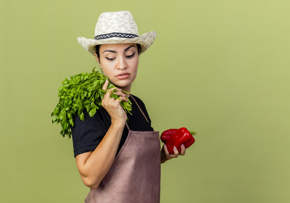 草药年轻漂亮的女园丁 围着围裙 戴着帽子 手里拿着红甜椒和新鲜的草药 严肃的脸站在淡绿色的墙上 一边看一边衣服帽子人