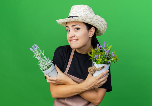 衣服年轻漂亮的女园丁 围着围裙 戴着帽子 手里拿着盆栽植物 站在绿色的墙边 微笑着困惑着抱着脸漂亮