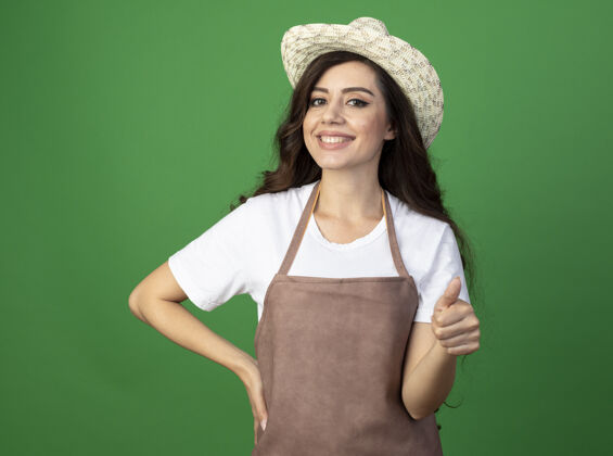 脸微笑的年轻女园丁穿着制服戴着园艺帽大拇指孤立地站在绿色的墙上年轻人人制服