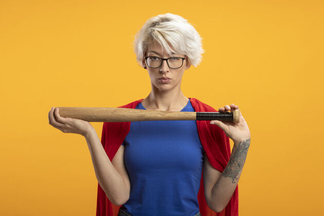 脸自信的女超人穿着红色斗篷 戴着光学眼镜 手持棒球棒 看着橙色墙壁上与世隔绝的前方眼镜超级英雄人
