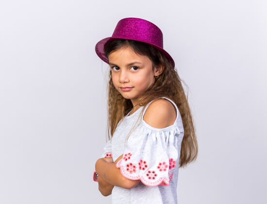 侧着可爱的白人小女孩 戴着紫色的派对帽 交叉着双臂 站在白色的墙上 留着复制空间帽子生日请