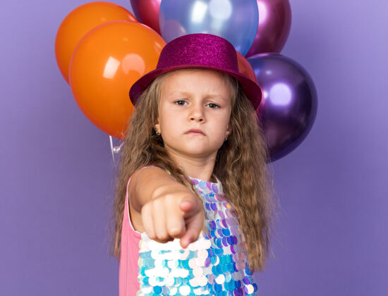 小严肃的金发小女孩 戴着紫色的派对帽 站在氦气球前 氦气球被隔离在紫色的墙上 留着复制空间生日站指向