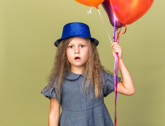 橄榄惊讶的金发小女孩 戴着蓝色派对帽 手里拿着氦气球 被隔离在橄榄绿的墙上 还有复制空间惊喜生日氦