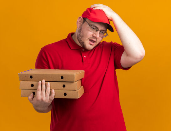头焦急的超重的年轻送货员戴着眼镜 手里拿着披萨盒 手放在头上 隔离在橙色的墙上 留着复印空间孤立焦虑年轻