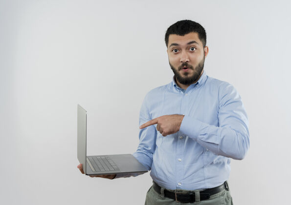 表情身穿蓝色衬衫 留着胡须的年轻人拿着笔记本电脑 用食指指着站在白墙上的笔记本电脑姿势人胡子