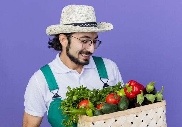 帽子年轻的留着胡须的园丁 穿着连体衣 戴着帽子 手里拿着装满蔬菜的箱子 站在蓝色的墙上开心地笑着表情年轻人脸