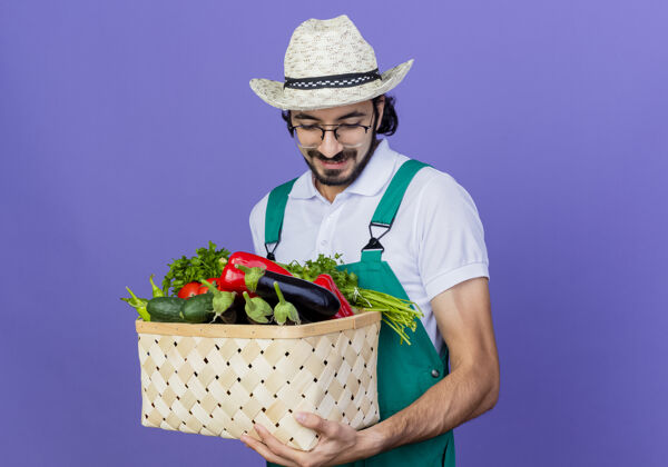 姿势年轻的留着胡须的园丁 穿着连体衣 戴着帽子 手里拿着装满蔬菜的箱子 站在蓝色的墙上微笑着看着下面感觉微笑蔬菜