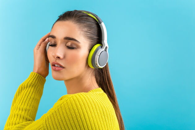 设备戴着无线耳机听音乐的女人开心地穿着黄色针织毛衣摆着蓝色的姿势装置声音姿势