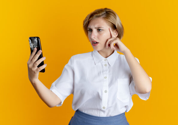 橙色年轻困惑的金发俄罗斯女孩把手放在脸上看电话金发电话背景