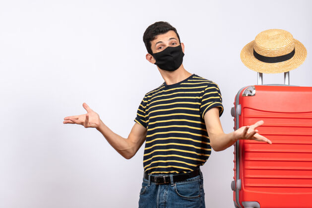 男人正面图：戴着黑色面具的年轻游客站在红色手提箱旁张开双手站立人面具
