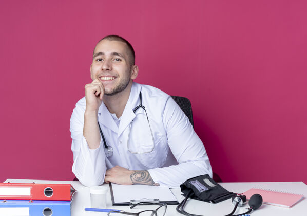 男性面带微笑的年轻男医生穿着医用长袍和听诊器坐在办公桌旁 手放在隔离在粉红色墙上的下巴上人医生表情
