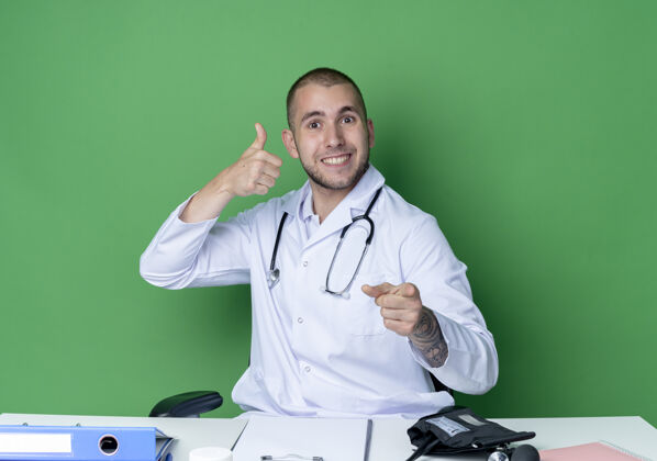 工具面带微笑的年轻男医生穿着医用长袍 听诊器坐在办公桌旁 工作工具向上伸出大拇指 指着隔离在绿色墙壁上的前方男性姿势工作