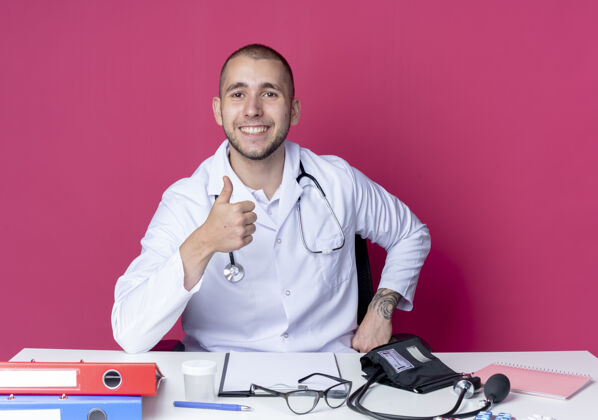 感觉面带微笑的年轻男医生穿着医用长袍 听诊器坐在办公桌旁 工作工具上的拇指被隔离在粉红色的墙上听诊器桌子粉色