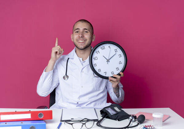 脸面带微笑的年轻男医生穿着医用长袍和听诊器坐在办公桌旁 手里拿着工作工具 把时钟和竖起的手指隔离在粉红色的墙上医生粉色人
