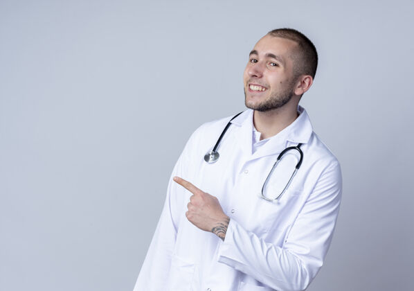 人面带微笑的年轻男医生 身穿医用长袍 脖子上戴着听诊器 指着隔离在白墙上的一侧男性脸人