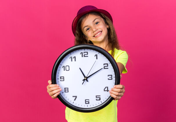 帽子可爱的白人小女孩 戴着紫色派对帽 把钟孤立地放在粉红色的墙上 留着复制空间钟小的女孩