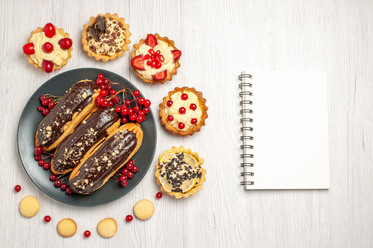 小吃俯瞰图：灰色盘子上的巧克力蛋糕和葡萄干 周围有馅饼和饼干 白色木桌上有笔记本 还有复印空间帽子食物薯片