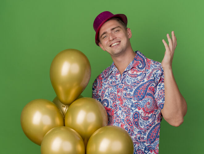 派对面带微笑的年轻人戴着粉红色的帽子拿着气球 举起手孤立地站在草地上气球年轻人举起