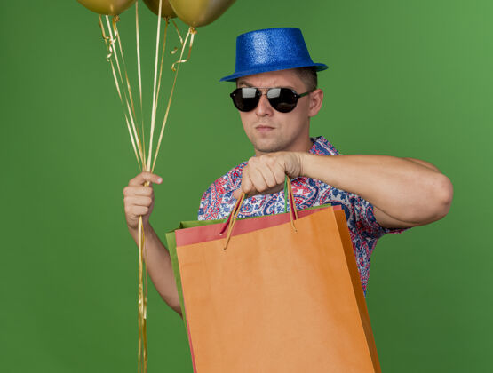 气球严格的年轻人戴着蓝色的帽子和眼镜拿着气球和礼品袋隔离在绿色眼镜帽子礼物