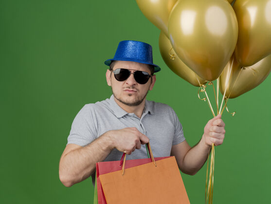 包严格的年轻人戴着聚会帽和眼镜拿着气球和礼品袋隔离在绿色眼镜戴着小伙子