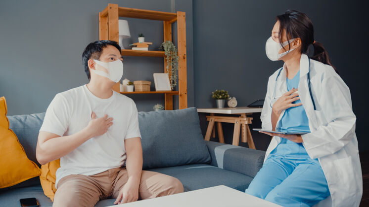 医疗保险年轻的亚洲女医生戴着口罩用数字平板电脑和快乐的男病人分享健康测试的好消息坐在家里的沙发上助理年轻人流行病