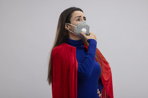 穿坚强的年轻超人女孩戴着医用口罩看着侧面 抓住下巴隔离在白色上思考壁板下巴