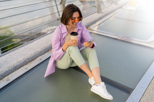 积极时尚短发女子坐在摩登桥上 一边喝咖啡一边用手机快乐漂亮女士
