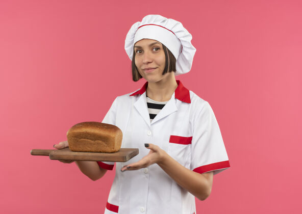 公民年轻的女厨师穿着厨师制服 手里拿着一块烤面包的砧板 隔离在粉红色的墙上持有厨师表情