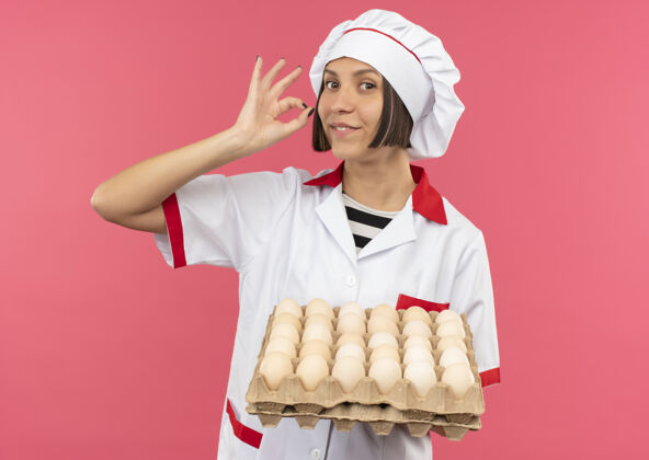 纸箱面带微笑的年轻女厨师身着厨师制服 手里拿着一盒鸡蛋 粉红色的墙上贴着“一切正常”的牌子人人制服