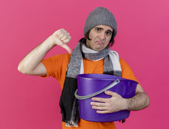 帽子悲伤的生病的年轻人戴着冬天的帽子 戴着围巾 拿着塑料桶 拇指朝下 孤立地站在粉红色的地板上水桶穿放下