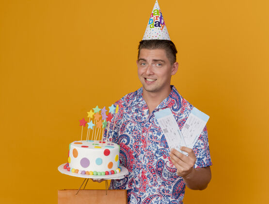 生日微笑的年轻人戴着生日帽 手里拿着生日礼物 蛋糕和票子孤立在橙色上橘子家伙举行