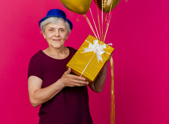 姿势面带微笑的老妇人戴着聚会帽 手里拿着氦气球和礼品盒 隔离在粉红色的墙上女人人老人
