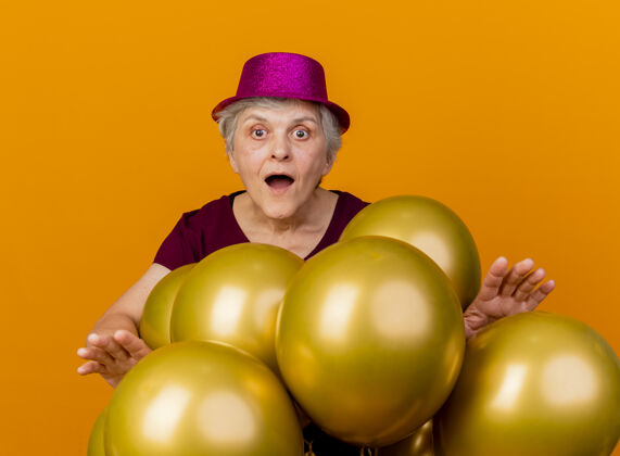 姿势惊讶的老妇人戴着派对帽站在氦气球面前 看着孤立的橙色墙壁帽子惊喜穿