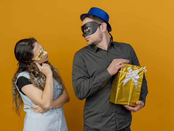 每个年轻的派对情侣互相看着戴着化装眼罩的家伙拿着礼盒孤立在橙色上眼睛橙色年轻人
