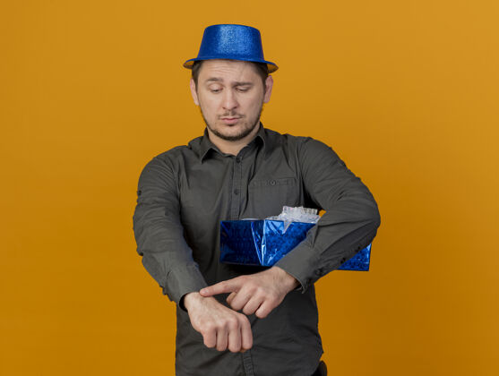 手势一个戴着蓝色帽子 拿着礼盒 展示着腕表的年轻人 在橙色的墙上孤立着时钟手腕盒子