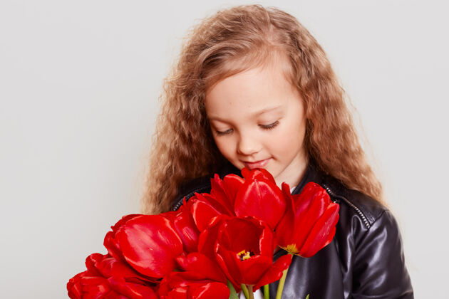 孩子金发可爱的小女孩手里捧着一束红色的郁金香 闻着美丽的花朵 低头 有着梦幻般的神情 穿着皮夹克肖像女学生乐观