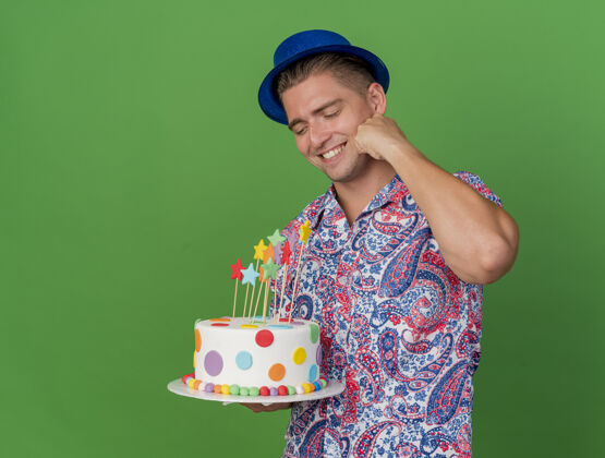 派对微笑着的年轻人戴着蓝色的帽子 拿着蛋糕 手放在脸上 隔着绿色的脸抱着绿色蓝色