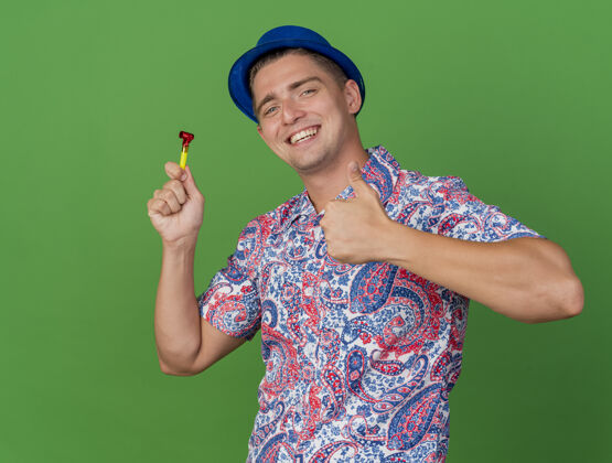 蓝色微笑着的年轻人戴着蓝色的帽子举行聚会吹风机显示拇指孤立的绿色年轻吹风机抱