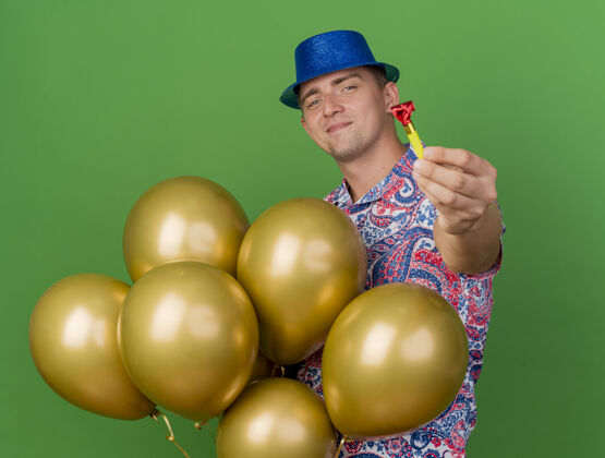 吹风机高兴的年轻人戴着蓝色的帽子 拿着气球 把聚会吹风机放在绿色的草地上手持年轻人绿色