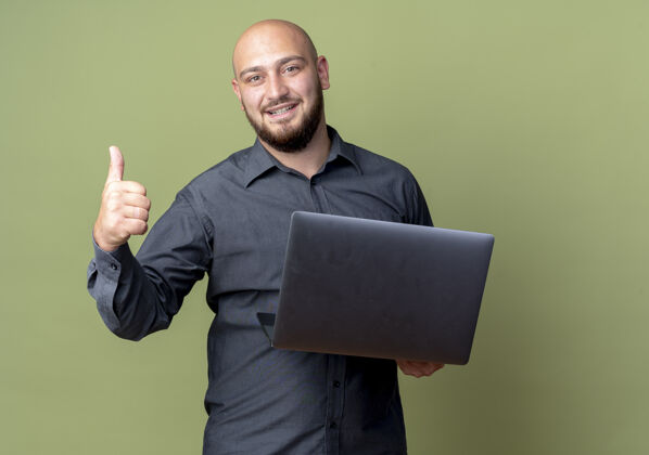 抱着微笑的年轻秃顶呼叫中心男子手持笔记本电脑 并显示拇指上孤立的橄榄绿墙上电话公民人
