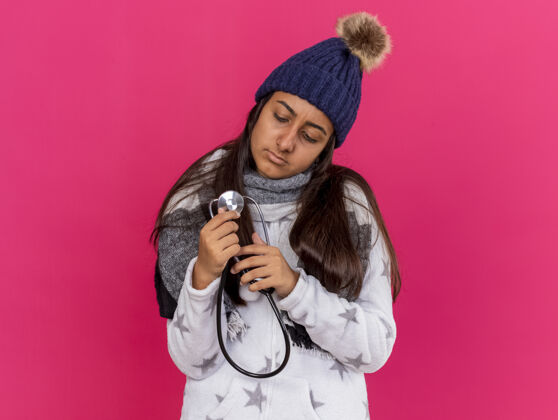 戴着生病的小女孩戴着冬天戴的帽子 戴着围巾 看着听诊器孤立地站在粉红色的墙上听诊器思考帽子