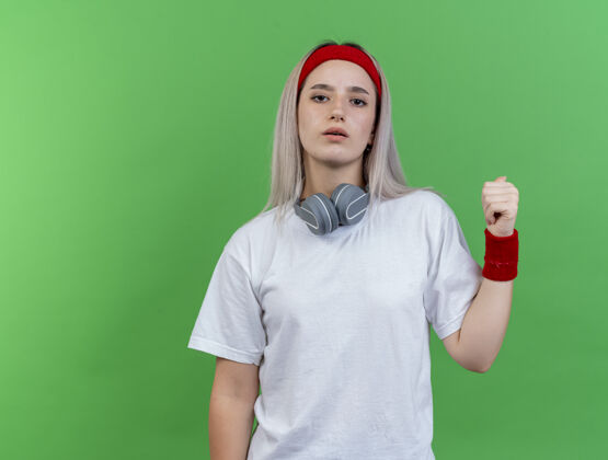 女人自信的年轻运动女性 戴着背带 戴着头带和腕带 脖子上戴着耳机 背对着绿色的墙壁耳机穿背部