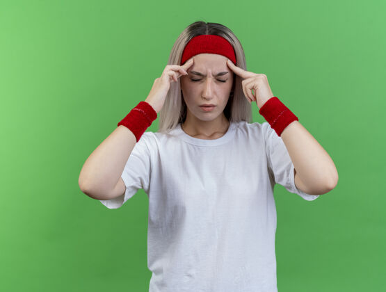 头带带着护腕戴着头带和腕带的年轻运动女性把手指放在隔离在绿色墙上的太阳穴上手指疼痛背带