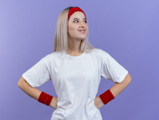 头带微笑着的年轻运动女性戴着背带 戴着头带和腕带 双手放在腰上 看着紫色墙上孤立的一面背带女孩腰