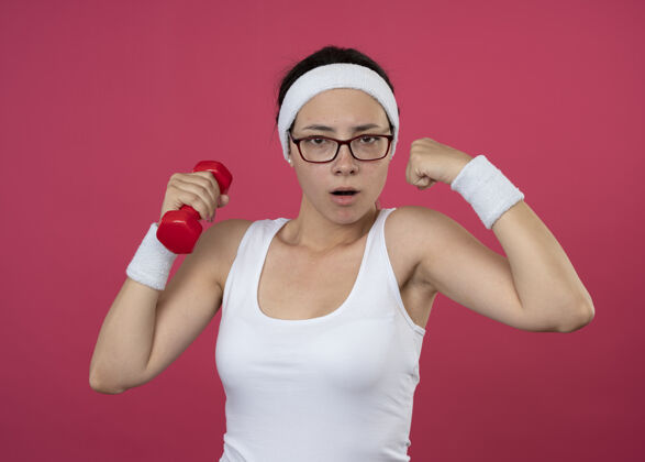 持有自信的年轻运动女性戴着眼镜戴着头带和腕带拿着哑铃和紧张的二头肌隔离在粉红色的墙上人自信年轻人