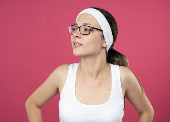 年轻戴着眼镜 戴着头带和腕带的快乐的年轻运动女性看着隔离在粉红色墙上的一面壁板女孩头带