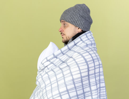 疾病苍白的年轻金发病男子戴着冬天的帽子和围巾包裹在格子布站在一旁举行的橄榄绿墙隔离枕头帽子裹着格子