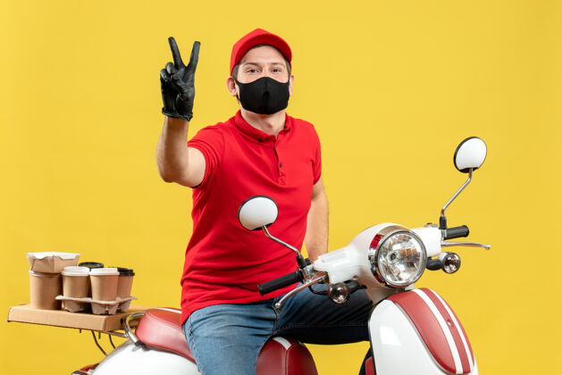 制造俯视图：微笑的快递员身穿红色上衣 戴着帽子手套 戴着医用面罩 坐在滑板车上做着胜利手势男性摩托车医疗
